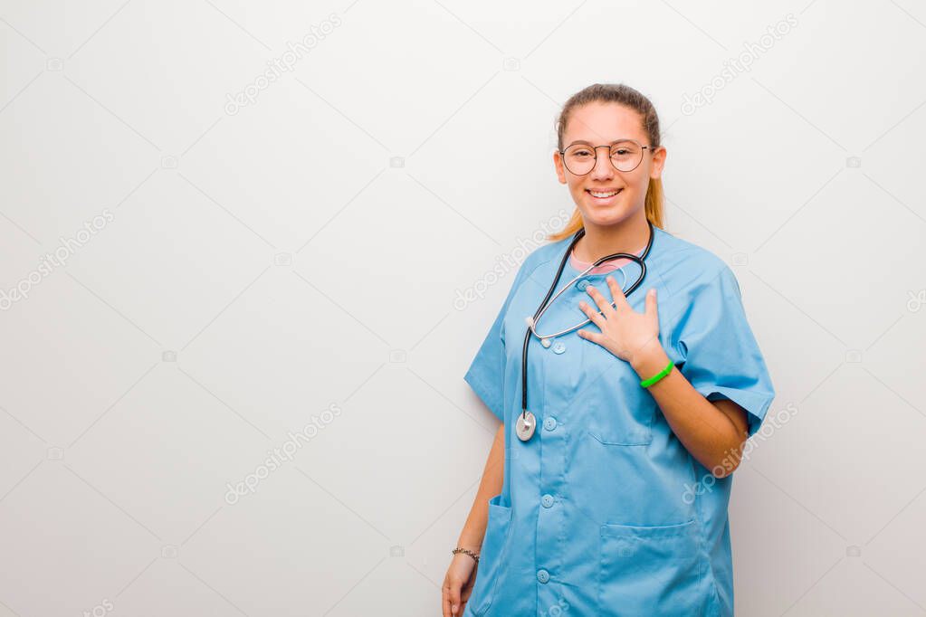 young latin nurse looking happy, surprised, proud and excited, pointing to self against white wall