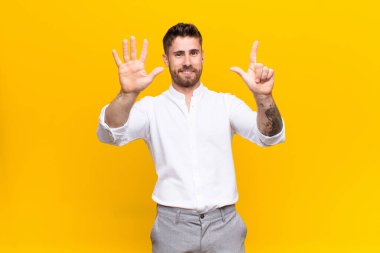 Genç adam gülümsüyor ve dost canlısı görünüyor, 7 ya da 7 numarayı gösteriyor, eli önde, düz renkli duvara doğru geri sayıyor.