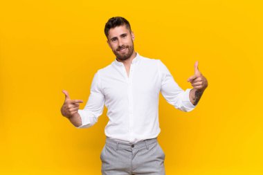 Genç adam gururlu, kibirli, mutlu, şaşırmış ve tatmin olmuş görünüyor, kendini gösteriyor, düz renkli duvara karşı kazanan gibi hissediyor.