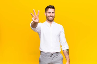 Genç adam gülümsüyor ve arkadaş canlısı görünüyor, üç ya da üç numarayı gösteriyor, düz renkli duvara doğru sayıyor.