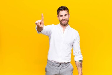 Genç adam gülümsüyor ve arkadaş canlısı görünüyor, bir numara ya da ilk eli önde, düz renkli duvara doğru sayıyor.