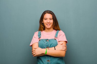 Genç, güzel, Latin bir kadın mutlu, gururlu ve tatmin olmuş görünüyor. Kollarını grunge duvarına dolamış gülümsüyor.