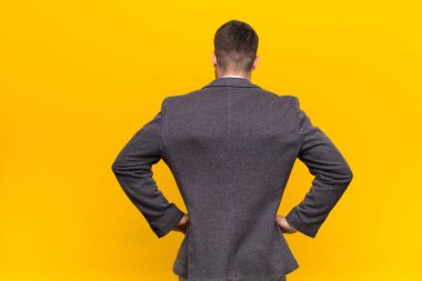 Genç handome erkeği kafası karışık ya da dolu hissediyor ya da şüphe ve sorular, merak ediyor, elleri kalçasında, düz renkli duvara karşı dikiz manzaralı