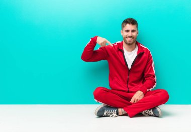 Genç adam mutlu, gururlu ve şaşırmış görünüyor, neşeli bir şekilde kendini gösteriyor, kendinden emin ve düz renkli duvara karşı yüce hissediyor.