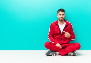 Genç adam gururlu, kendinden emin ve mutlu görünüyor, gülümsüyor ve kendini işaret ediyor ya da düz renk duvara karşı bir numara işareti yapıyor.