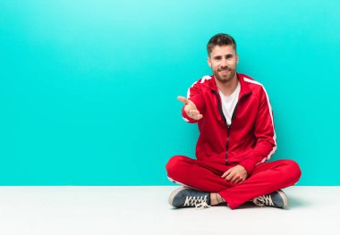Genç adam gülümsüyor, mutlu, kendinden emin ve arkadaş canlısı görünüyor, anlaşmayı bitirmek için el sıkışmayı teklif ediyor, düz renkli duvara karşı işbirliği yapıyor.