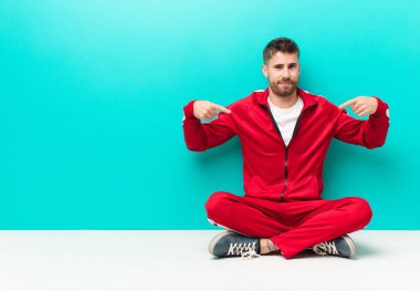 Genç adam gururlu, pozitif ve rahat görünüyor. İki elini de düz duvara dayayıp göğsünü gösteriyor.