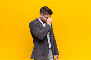 Genç adam stresli, mutsuz ve hayal kırıklığına uğramış, alnına dokunmuş ve düz renkli duvara karşı şiddetli baş ağrısı çeken bir adam.