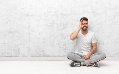 Genç adam mutlu bir şekilde gülümsüyor, şaka yapıyor ve gözetleme deliğinden bakıyor, düz renkli duvara karşı sırları gözetliyor.