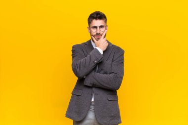 Genç adam ciddi, düşünceli ve güvensiz görünüyor, bir kolu çapraz ve çene üzerinde, düz renk duvarla seçenekleri tartıyor