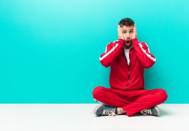 Genç handosme adam mutsuz, korkmuş ya da endişeli görünüyor, ağzı sonuna kadar açık ve iki kulağı da düz renkli duvara karşı kapatıyor.