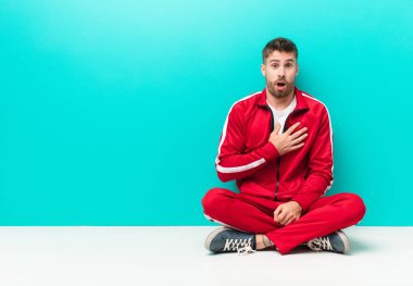 Genç adam şok olmuş, şaşırmış ve şaşırmış hissediyor, elleri göğsünde ve ağzı açık, kim diyor, ben mi? yassı renk duvarına karşı