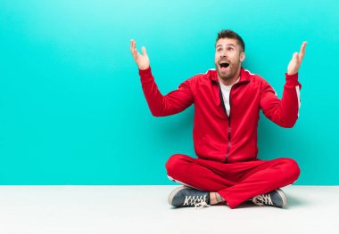 Genç adam kendini mutlu, şaşkın, şanslı ve şaşırmış hissediyor. İki eli havada, düz renkli duvara karşı zaferi kutluyor.