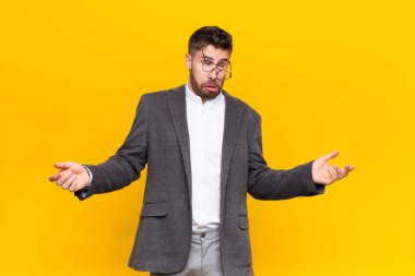 Genç adam aptal, çılgın, kafası karışık, kafası karışmış bir ifadeyle omuz silkiyor. Sinirli ve düz renkli duvara karşı cahil hissediyor.