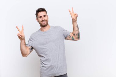 Genç adam gülümsüyor ve mutlu görünüyor, arkadaş canlısı ve memnun, iki eliyle düz duvara karşı zafer ya da barış işareti yapıyor