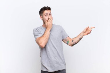 Genç adam kendini mutlu, şaşkın ve şaşırmış hissediyor. Ağzını eliyle kapatıyor ve düz renkli duvara karşı yanal fotokopi alanını işaret ediyor.