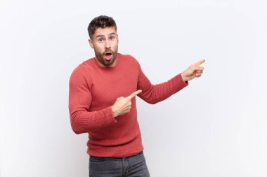 Genç adam şok olmuş ve şaşırmış hissediyor, yan taraftaki boşluğu işaret ederek, düz renk duvara karşı şaşkın, açık ağızlı bir görünüm