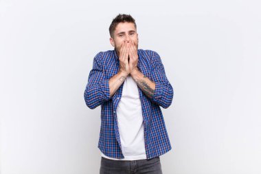 Genç adam mutlu ve heyecanlı, şaşırmış ve şaşırmış elleriyle ağzını kapatmış, düz renkli duvara karşı sevimli bir ifadeyle kıkırdıyor.