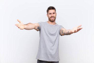 Genç adam neşeyle gülümsüyor sıcak, dostça, sevgi dolu bir hoş geldin sarılması yapıyor, mutlu ve sevimli hissediyor düz renkli duvara karşı.