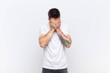 Genç adam üzgün, hayal kırıklığına uğramış, gergin ve depresif hissediyor, iki eliyle yüzünü kapatıyor, düz renkli duvara karşı ağlıyor.