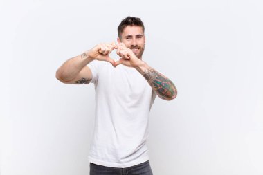Genç adam gülümsüyor ve mutlu, sevimli, romantik ve aşık, iki eliyle düz renkli duvara kalp şekli veriyor.