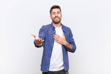 Genç adam mutlu ve aşık hissediyor, bir eli kalbinin yanında gülümsüyor ve diğeri düz renkli duvara doğru uzanıyor.