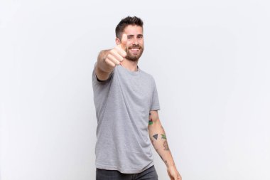 Genç adam gururlu, kaygısız, kendinden emin ve mutlu hissediyor. Düz renkli duvara yaslanmış olumlu gülümsüyor.