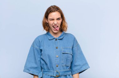 Genç sarışın kadın neşeli, kaygısız, asi tavırlı, şakacı ve dilsiz, düz renkli duvara karşı eğlenen