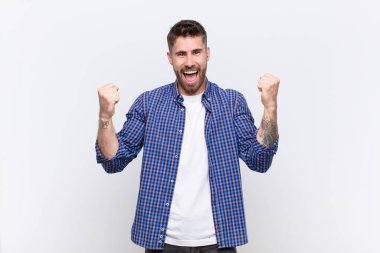 Genç adam mutlu, şaşırmış ve gururlu hissediyor, bağırıyor ve başarıyı kutluyor. Düz renkli duvara karşı büyük bir gülümsemeyle.