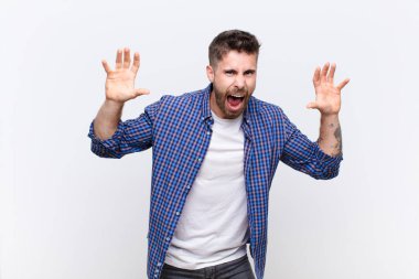 Genç handosme adam panik ya da öfke içinde bağırıyor, şok olmuş, korkmuş ya da öfkeli, elleri düz renkli duvara dayalı.