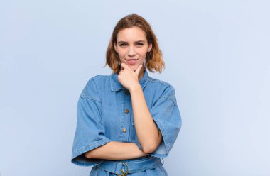 Genç, sarışın bir kadın mutlu, kendinden emin bir ifadeyle gülümsüyor. Elleri çenesinde, merak ediyor ve düz renkli duvara bakıyor.