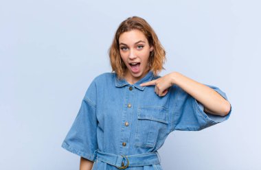 Genç sarışın kadın mutlu, gururlu ve şaşırmış görünüyor, neşeli bir şekilde kendini gösteriyor, kendinden emin ve düz renkli duvara karşı yüce hissediyor.