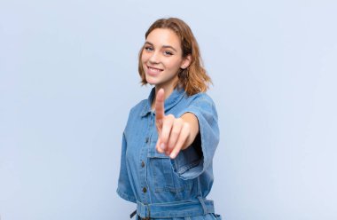 Genç sarışın kadın gururla gülümsüyor ve gururla bir numaralı pozunu veriyor. Düz renkli duvara karşı bir lider gibi hissediyor.