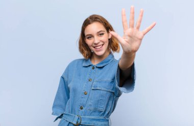 Genç sarışın kadın gülümsüyor ve arkadaş canlısı görünüyor, 5 ya da 5. numarayı gösteriyor, düz renkli duvara doğru sayıyor.