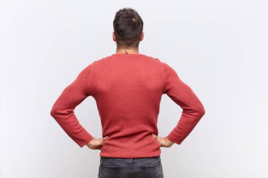 Genç handome erkeği kafası karışık ya da dolu hissediyor ya da şüphe ve sorular, merak ediyor, elleri kalçasında, düz renkli duvara karşı dikiz manzaralı