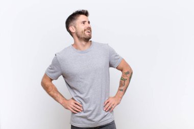 Genç adam mutlu, neşeli ve kendinden emin görünüyor. Gururla gülümsüyor ve iki elini de düz duvara yaslamış kalçaları kolluyor.