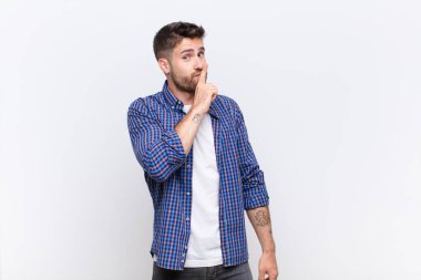 Genç handosme adam sessizlik ve sessizlik istiyor, parmaklarıyla ağzının önünde işaret ediyor, sessiz ol diyor ya da düz renkli duvara karşı bir sır saklıyor.