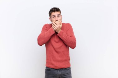Genç handosme adam şok olmuş, şaşırmış bir ifadeyle ağzını kapatıyor, bir sır saklıyor ya da düz renkli duvara karşı 