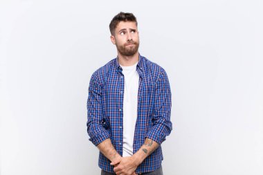 Genç adam şaşkın ve şaşkın görünüyor, merak ediyor ya da bir sorunu çözmeye çalışıyor ya da düz renkli duvara karşı düşünüyor.