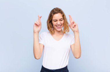 Genç sarışın kadın gergin ve umutlu, dua ediyor ve düz renkli duvara karşı şans diliyor.