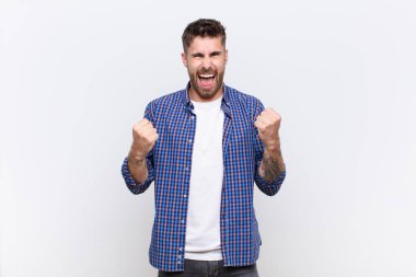 Genç handome 'lu muzaffer bir şekilde bağırıyor, gülüyor, mutlu ve heyecanlı hissederken düz renkli duvara karşı başarıyı kutluyor.