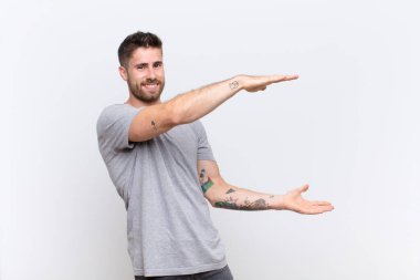 Genç adam gülümsüyor, mutlu, pozitif ve tatmin olmuş hissediyor, düz renkli duvara karşı fotokopi alanında nesne ya da konsept tutuyor ya da gösteriyor