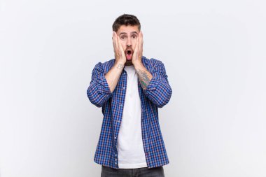 Genç adam şok olmuş ve korkmuş hissediyor, ağzı açık ve elleri yanaklarında düz renkli duvara bakıyor.