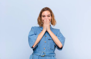 Genç sarışın kadın mutlu, neşeli, şanslı ve şaşırmış görünüyor. İki elini de düz duvara dayayıp ağzını kapatmış.