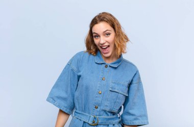 Genç, sarışın bir kadın, büyük, dost canlısı, kaygısız bir gülümseme, pozitif görünüyor, rahat ve mutlu, düz renkli duvara karşı ürpertici