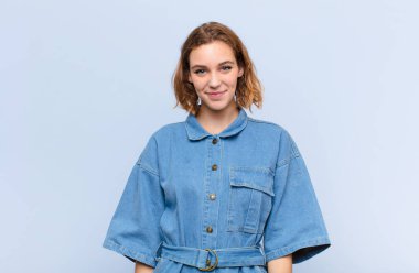 Genç sarışın kadın pozitif ve kendinden emin gülümsüyor. Tatmin olmuş, arkadaş canlısı ve mutlu görünüyor. Düz renkli duvara karşı.