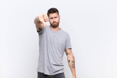 Huysuz, sinirli, sinirli, hayal kırıklığına uğramış ya da canı sıkkın genç bir adam, düz renk duvarına karşı ciddi bir bakışla başparmaklarını aşağı gösteriyor.