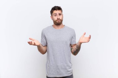 Genç adam şaşkın ve şaşkın hissediyor, şüpheci, farklı seçenekleri tartıyor ya da seçiyor düz renkli duvara karşı komik bir ifadeyle