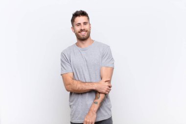 Genç adam utangaç ve neşeli bir şekilde gülüyor. Düz renkli duvara karşı dostane, olumlu ama güvensiz bir tutum sergiliyor.