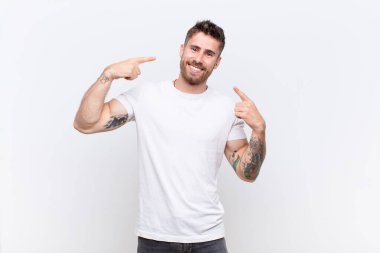 Genç adam kendine güvenerek gülümsüyor. Geniş bir gülümseme, pozitif, rahat, düz renkli duvara karşı tatmin olmuş bir tavır.